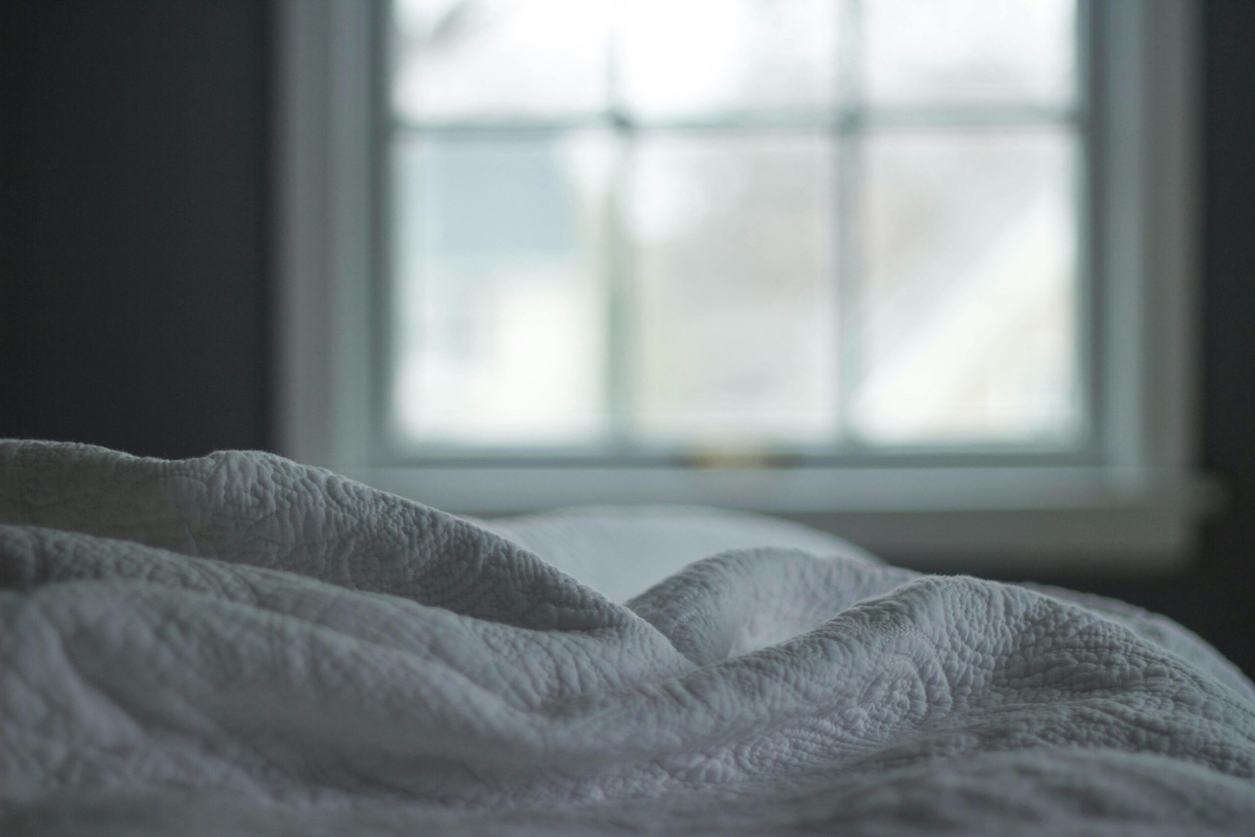 Image of a bed with white sheets and a blurred window in the background.